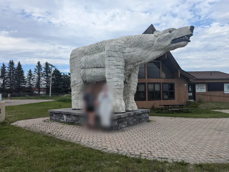 A giant carved white polar bear statue on a paving-stone plinth outside the Cochrane Board of Trade tourism building; two visitors in front of it are blurred.