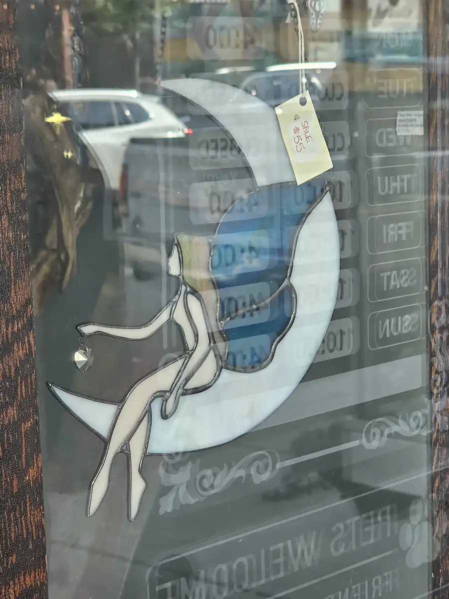 A stained-glass fairy sitting on a crescent moon, hanging in a downtown shop window with the street reflected in the glass.