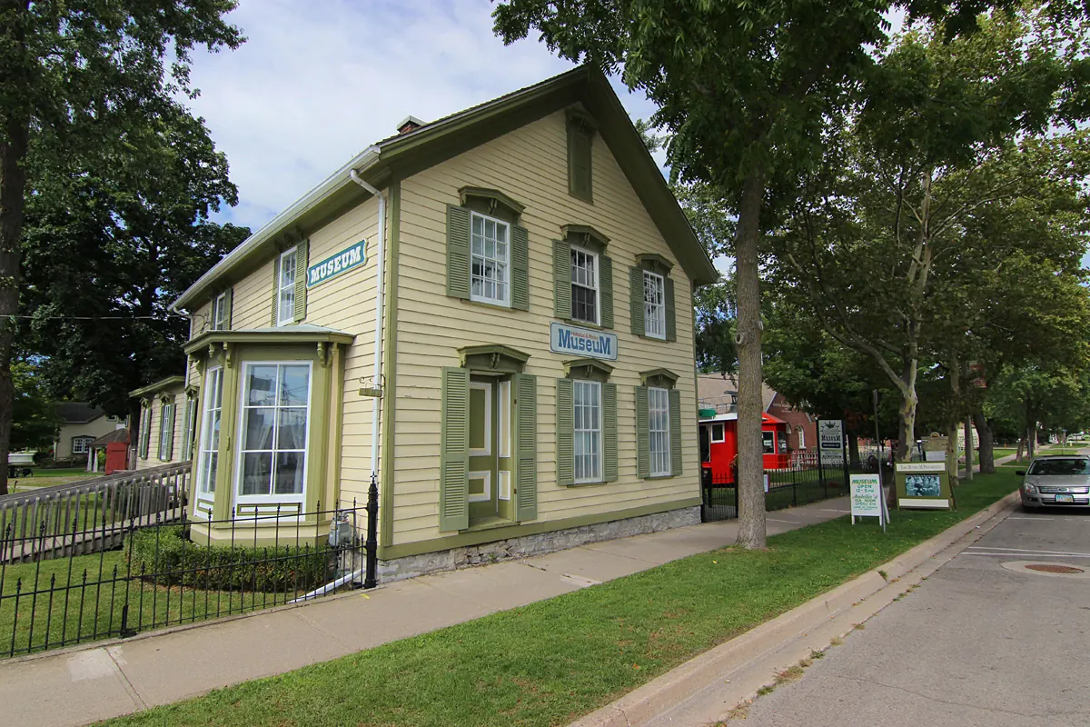 The Port Colborne Historical and Marine Museum, a pale green wooden house with a fenced garden on a tree-lined street.